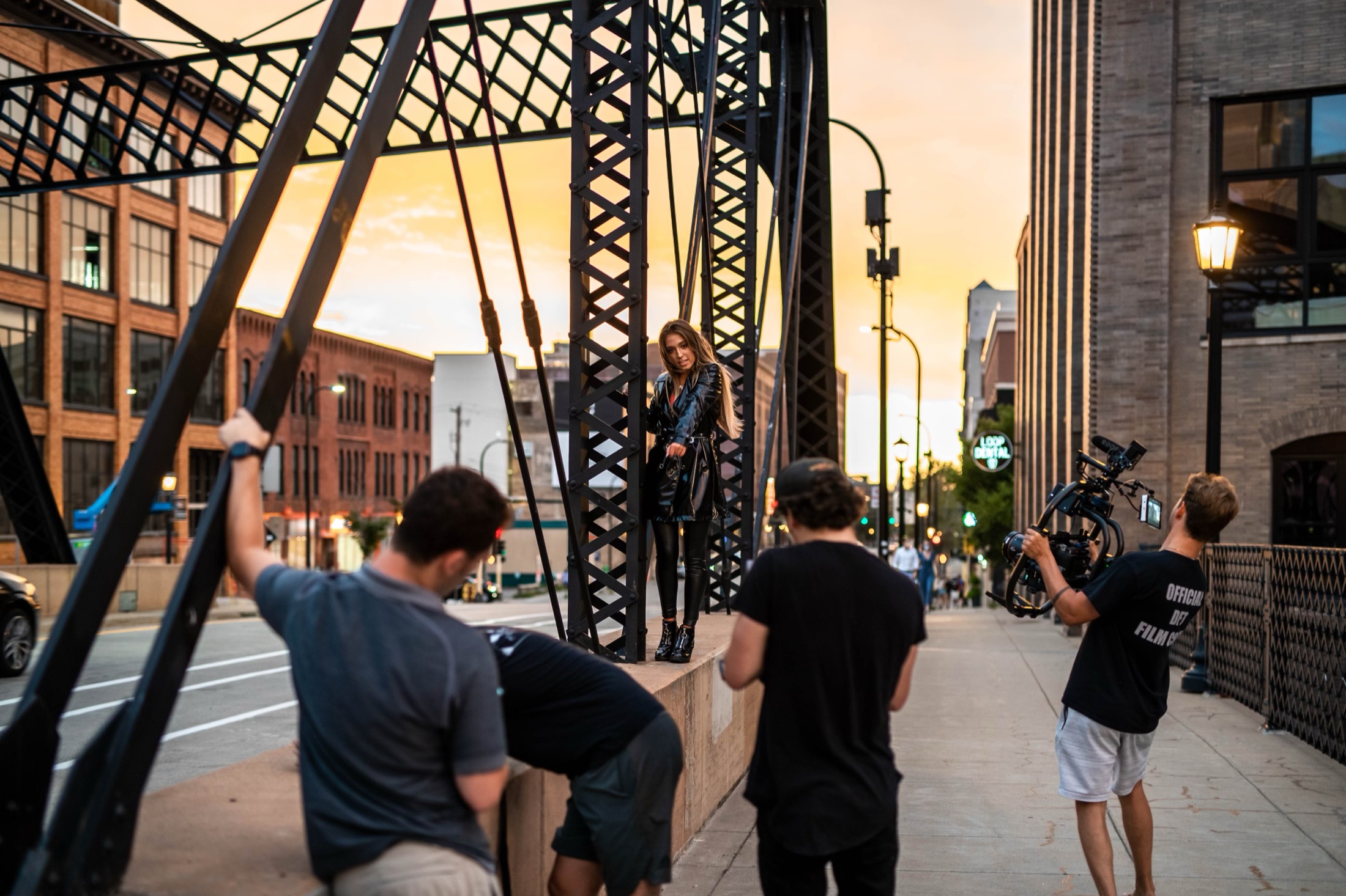 On-set camera team filming a talent on an urban bridge at sunset.