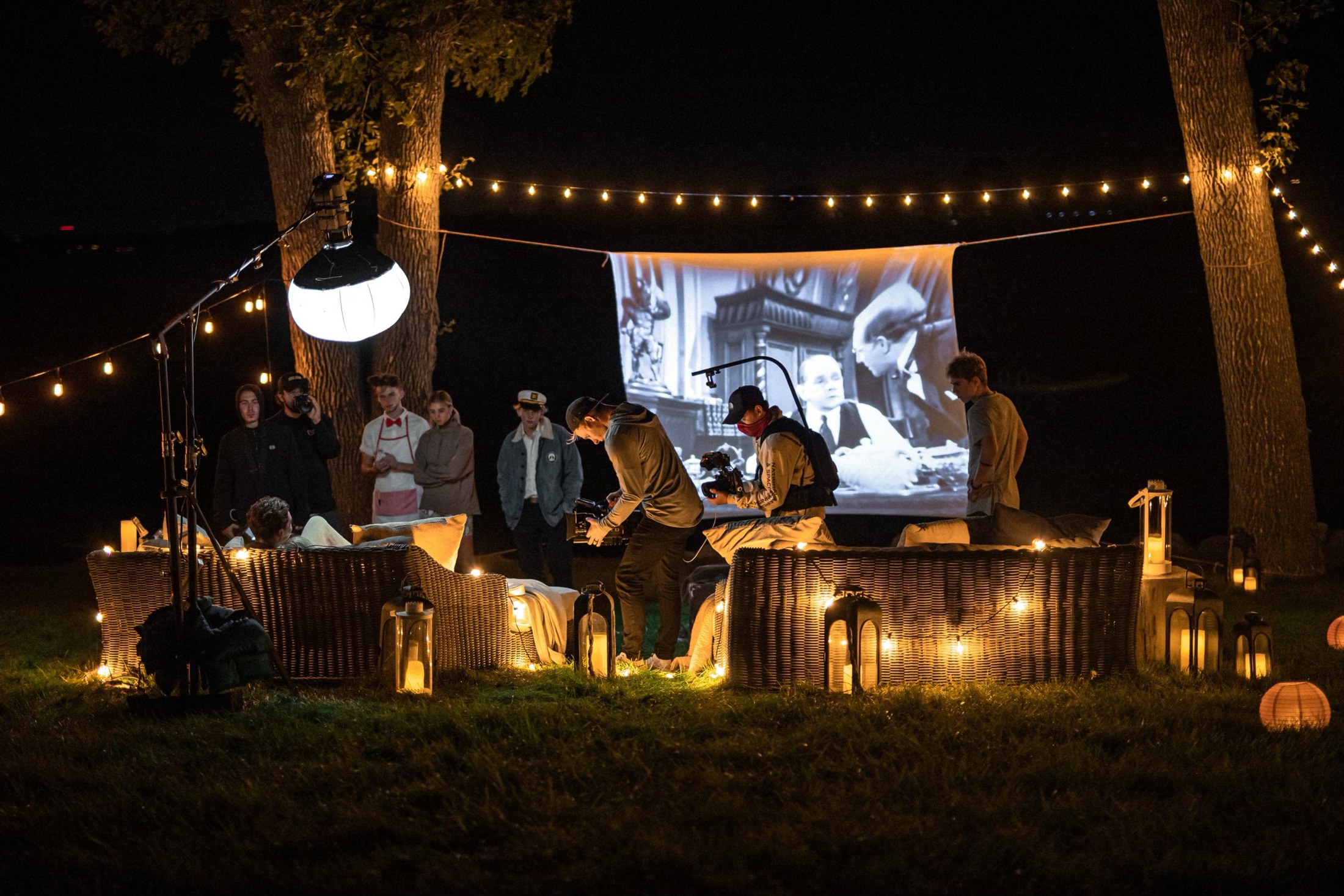 Night shoot with a projection screen, string lights, and camera crew.